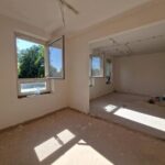 Pomieszczenie w remoncie o białych ścianach, z otwartymi oknami. Podłoga z plamami farby, materiałami i sprzętem. W tle widać zielone drzewa.