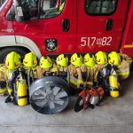 Na zdjęciu widać ośmiu strażaków w żółtych hełmach i brązowych mundurach, stojących przed czerwonym wozem strażackim. Na ziemi leżą sprzęty: wentylator i przecinarka. Tło stanowi wnętrze garażu.