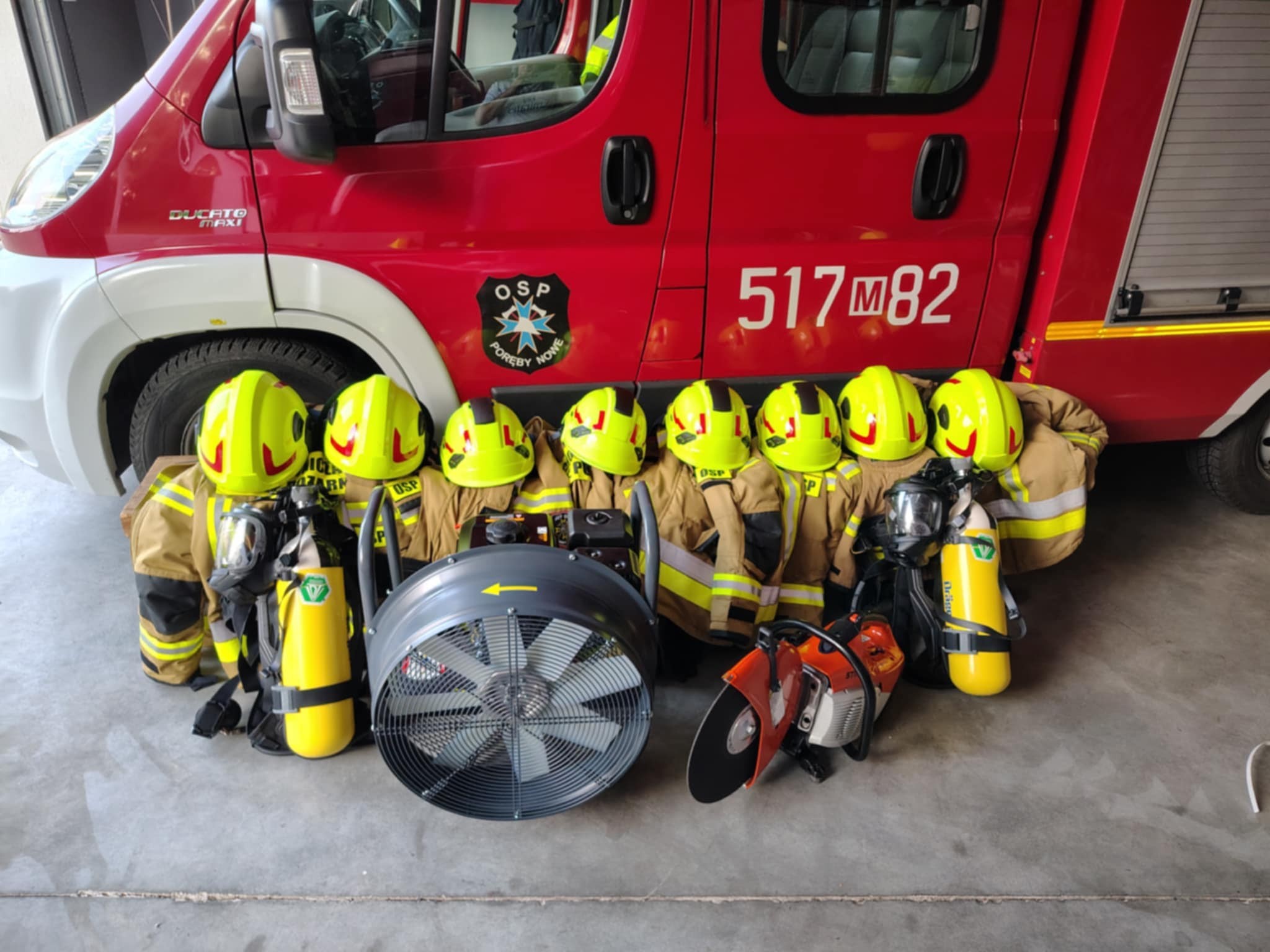 Na zdjęciu widać ośmiu strażaków w żółtych hełmach i brązowych mundurach, stojących przed czerwonym wozem strażackim. Na ziemi leżą sprzęty: wentylator i przecinarka. Tło stanowi wnętrze garażu.