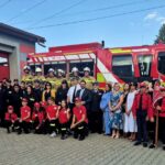 Grupa strażaków i osób w kolorowych mundurach stoi przed czerwoną remizą i wozem strażackim na tle drzew.