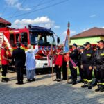 Czerwony wóz strażacki otoczony strażakami w strojach galowych stoi przed budynkiem. W centralnej części ksiądz dokonuje poświęcenia pojazdu. Na niebie są białe chmury.