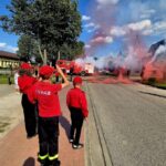 Grupa ludzi w czerwonych strojach strażackich stoi na chodniku. Na ulicy unoszą się kolorowe dymy, a w tle widać czerwony wóz strażacki. Pogodne niebo i drzewa tworzą tło.