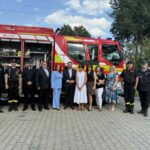 Grupa ludzi stoi przed czerwonym wozem strażackim na tle zielonych drzew i błękitnego nieba. Widać strażaków i osoby w eleganckich ubraniach. Tło jest jasne i słoneczne.