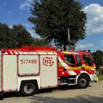 Wóz strażacki na parkingu, czerwono-biały z numerem 112. W tle drzewa i błękitne niebo. Z boku widoczne ludzię.