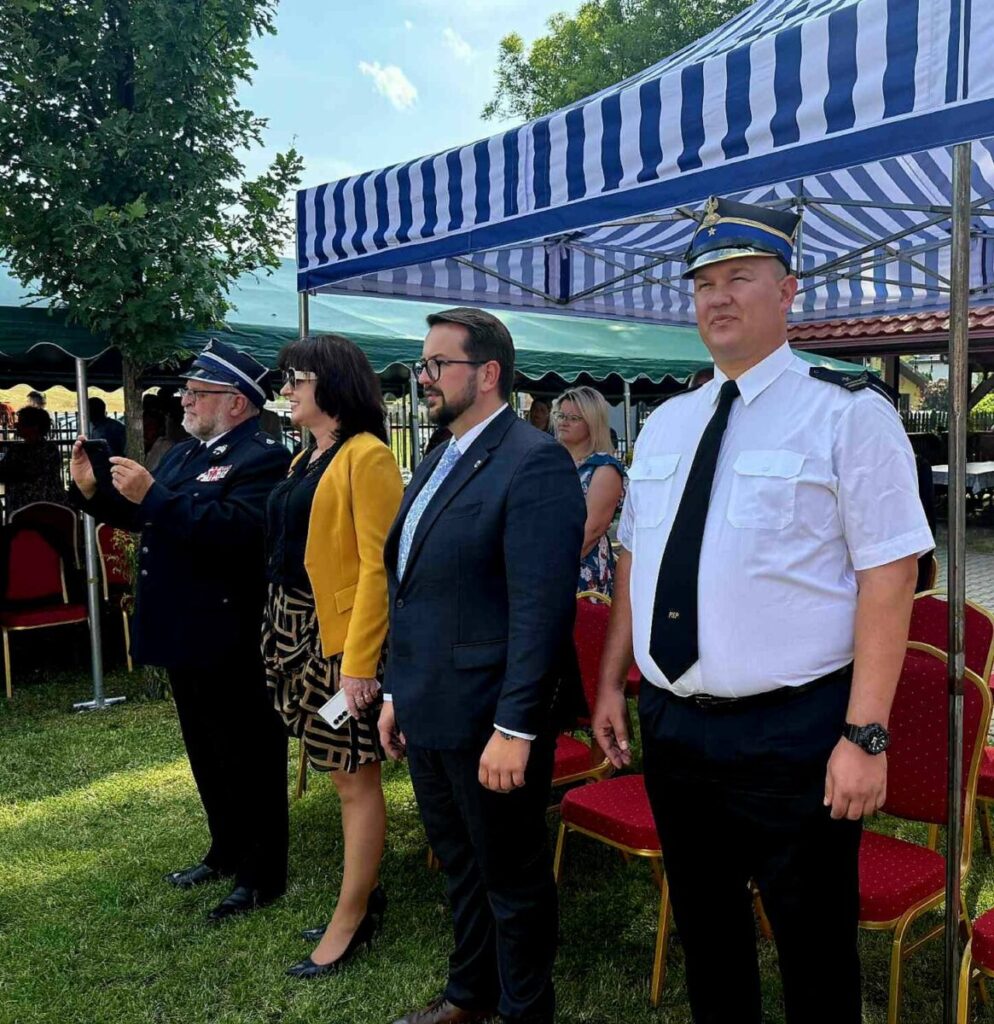 Grupa osób stoi na trawie pod niebiesko-białym namiotem. Mężczyźni i kobieta ubrani są formalnie, z tyłu widać drzewa i krzesła. Jedna osoba ma na sobie mundur.