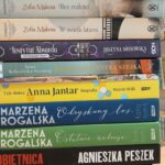 Stos książek o różnorodnych okładkach, od czarnej przez pomarańczową po niebieską, stoi na brązowym tle. Różne tytuły i autorzy na grzbietach. Kolorowe napisy wyróżniają poszczególne tytuły.