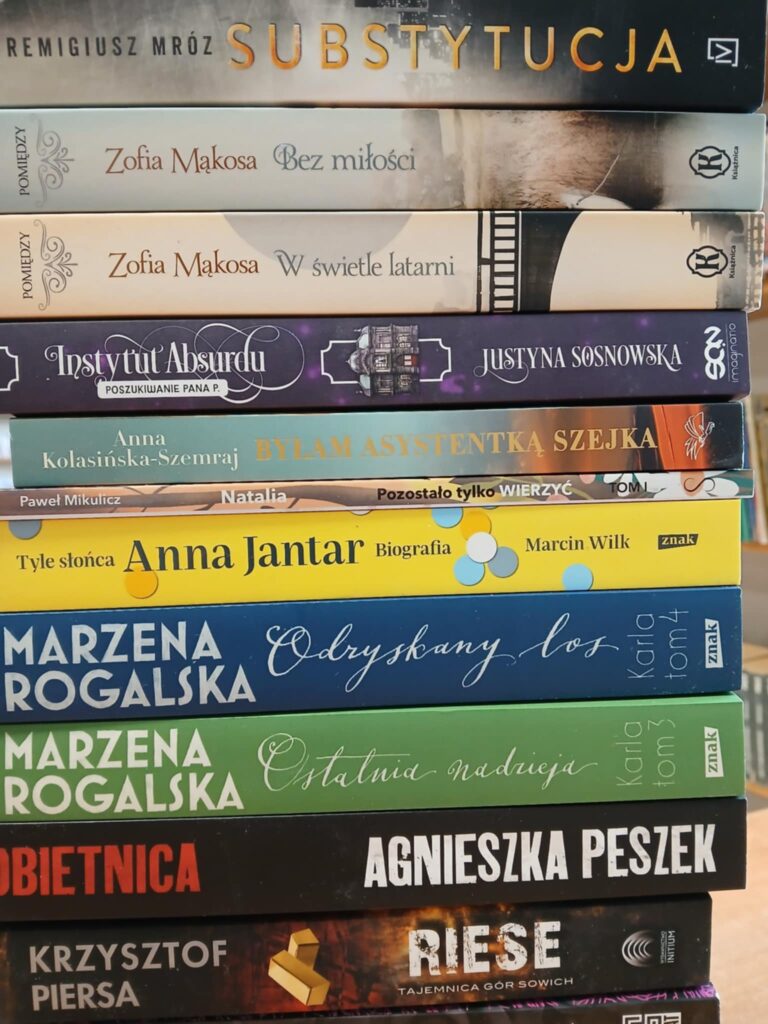 Stos książek o różnorodnych okładkach, od czarnej przez pomarańczową po niebieską, stoi na brązowym tle. Różne tytuły i autorzy na grzbietach. Kolorowe napisy wyróżniają poszczególne tytuły.