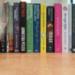 Rząd książek na półce z różnorodnymi okładkami. Kolory dominujące to czarny, zielony, biały i różowy. Tło to drewniana powierzchnia.