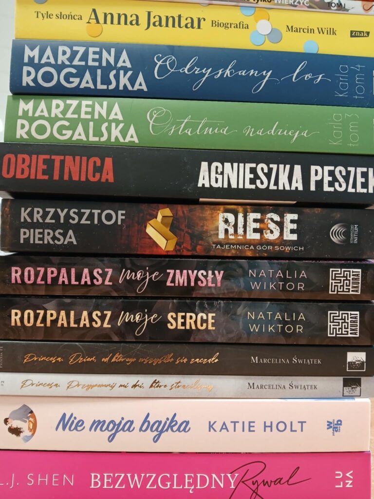 Stos książek z różnobarwnymi grzbietami, od żółci, przez zieleń, po róż. Każda książka ma widoczny tytuł i autora. Tło jest białe.