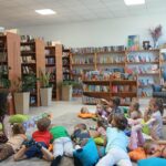 548173565_1348118723657728_1134729478398374430_n Grupa dzieci leży na poduszkach w bibliotece, słuchając dorosłej osoby. Tło stanowią drewniane regały pełne książek. Kolorowe poduszki i zabawki tworzą przytulną atmosferę.