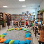 548891465_1076100717906678_5187707014709542699_n Grupa dzieci bawi się w sali z drewnianymi półkami pełnymi książek. Na podłodze leżą kolorowe poduszki. Ściany są jasne, a wnętrze jest dobrze oświetlone.