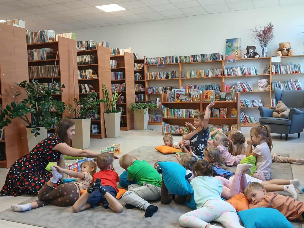 Grupa dzieci leży na poduszkach w kolorowej bibliotece, słuchając kobiety. W tle półki z książkami, rośliny i pluszowe misie. Na podłodze jasny dywan.