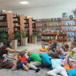 549098891_1076101151239968_5104968691502470434_n Grupa dzieci leży na poduszkach w kolorowej bibliotece, słuchając kobiety. W tle półki z książkami, rośliny i pluszowe misie. Na podłodze jasny dywan.