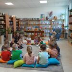 549155666_1076101061239977_1960240804207435290_n Grupa dzieci siedzi na podłodze w bibliotece na kolorowych poduszkach. W tle regały z książkami i rośliny. Prowadząca kobieta uczestniczy w zajęciach.
