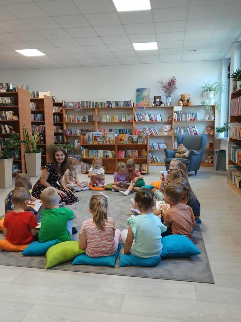 Grupa dzieci siedzi na podłodze w bibliotece na kolorowych poduszkach. W tle regały z książkami i rośliny. Prowadząca kobieta uczestniczy w zajęciach.