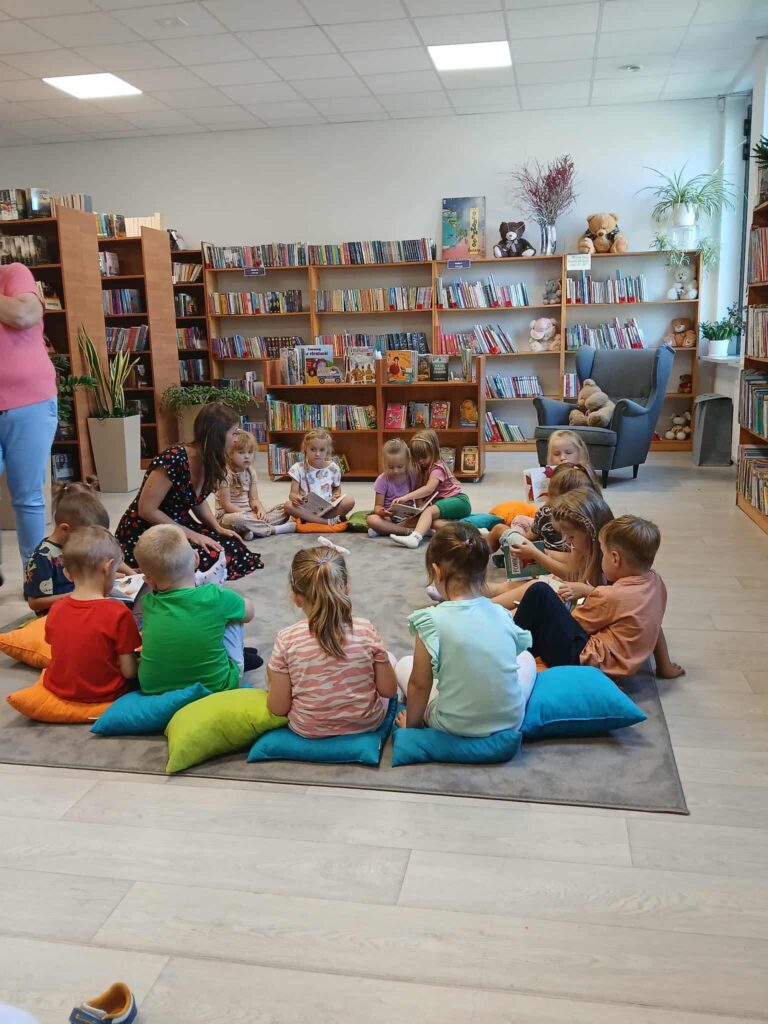 Dzieci siedzą na kolorowych poduszkach w bibliotece, zajęte książkami. Tło tworzą półki pełne książek, ściany jasne. Dookoła rośliny i maskotki dodają przytulności.
