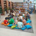 549729587_1076100587906691_8883358172852682330_n Dzieci siedzą na kolorowych poduszkach w jasnej bibliotece. Tło to półki z książkami i pluszowe misie. Rośliny dodają zieleni.