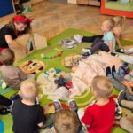 549797004_1076100987906651_231068132656518280_n Dzieci siedzą w kręgu na kolorowym dywanie w przedszkolu. Kobieta z chustką na głowie pokazuje im pacynki. W tle są szafki i zabawki.