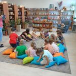 550166599_1076100861239997_6979118348035884263_n Grupa dzieci siedzi na kolorowych poduszkach w bibliotece. Na półkach w tle są książki i pluszaki. Oświetlenie jest jasne.