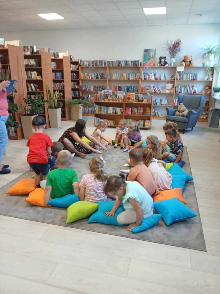 Grupa dzieci siedzi na kolorowych poduszkach w bibliotece. Na półkach w tle są książki i pluszaki. Oświetlenie jest jasne.