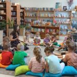 550658759_1076101097906640_1565039492939638257_n Grupa dzieci siedzi na kolorowych poduszkach w bibliotece. Przed nimi młoda kobieta czyta książkę. W tle regały z kolorowymi książkami.