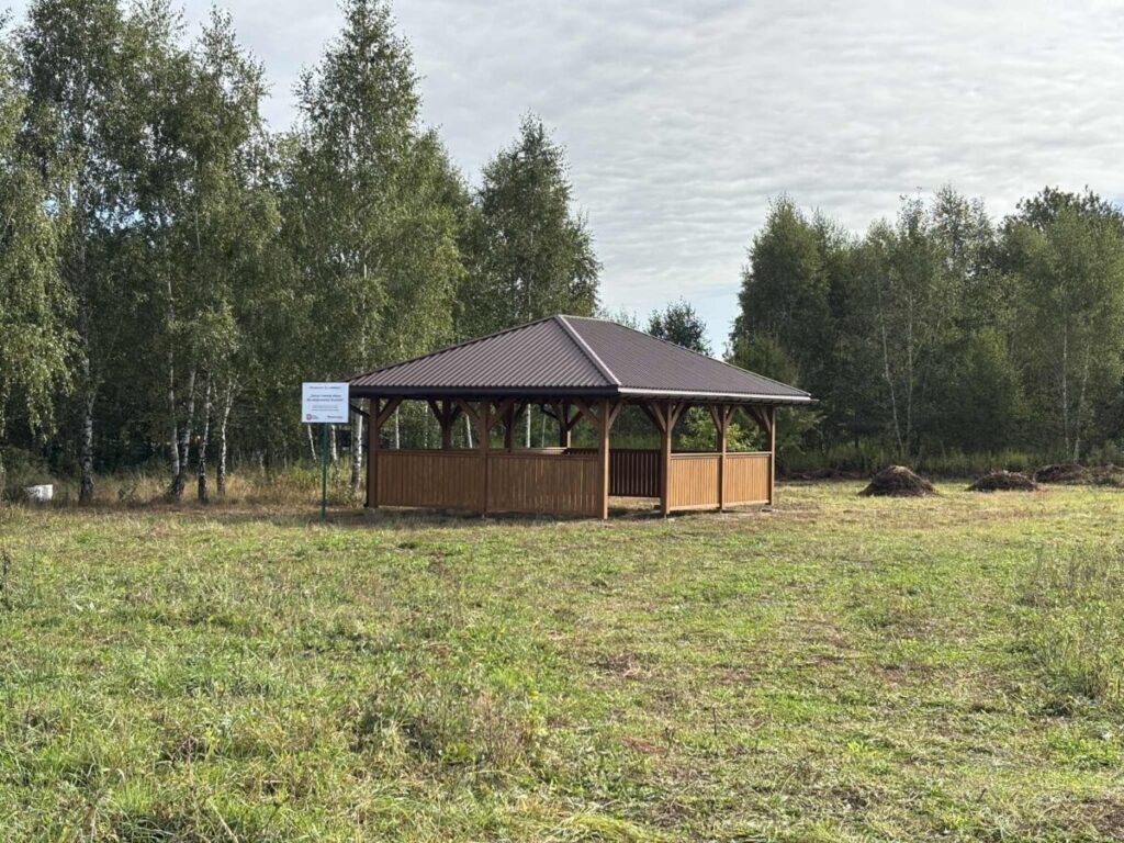 Drewniana altana z brązowym dachem stoi na tle zielonego lasu. Trawa na pierwszym planie jest niska. Obok altany znajduje się znak.