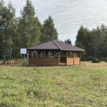 Drewniana altana z brązowym dachem stoi na tle zielonego lasu. Trawa na pierwszym planie jest niska. Obok altany znajduje się znak.