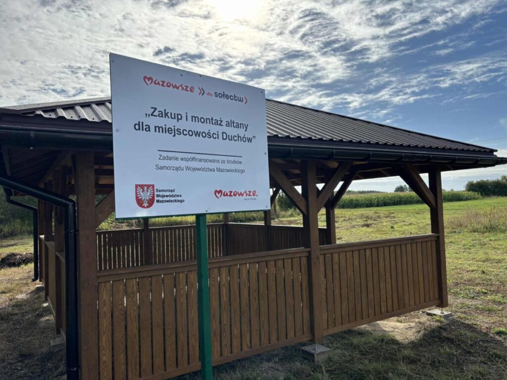 Drewniana altana na tle zielonego pola i błękitnego nieba. Na pierwszym planie biała tablica z czerwonym napisem. Altana ma ciemny dach.