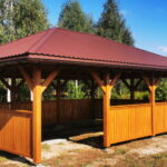 Drewniana altana z brązowym dachem stoi na trawie. Otoczona jest drzewami w tle. Jasne, słoneczne niebo dodaje kontrastu.