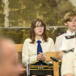 Sala z muzykami w mundurach; dziewczyna z trąbką siedzi z przodu. Tło w kolorach ziemi; kilka osób rozmytych.