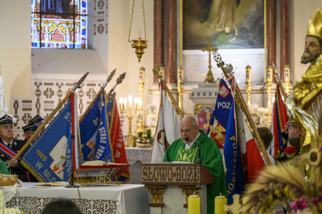 Mężczyzna w zielonej szacie przemawia w kościele, otoczony sztandarami. W tle witraż i świeczniki. Po prawej figura świętego.