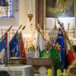 Mężczyzna w zielonej szacie przemawia w kościele, otoczony sztandarami. W tle witraż i świeczniki. Po prawej figura świętego.