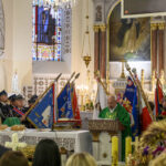 Wnętrze kościoła z duchownym przy ołtarzu. Kolorowe witraże i sztandary strażackie w tle. Złota figura świętego z prawej strony.