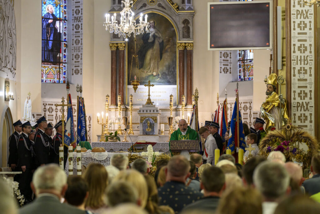 Wnętrze kościoła z ołtarzem, księdzem w zieleni i zgromadzeniem. Okna witrażowe i dekoracje w tle. Ludzie w ławkach obserwują ceremonię.