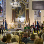 Wnętrze kościoła z ołtarzem, księdzem w zieleni i zgromadzeniem. Okna witrażowe i dekoracje w tle. Ludzie w ławkach obserwują ceremonię.