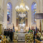 Wnętrze kościoła z ozdobnym ołtarzem i witrażami w tle. Jasne, złote żyrandole zwisają z sufitu. Ludzie w strojach galowych stoją przed ołtarzem.