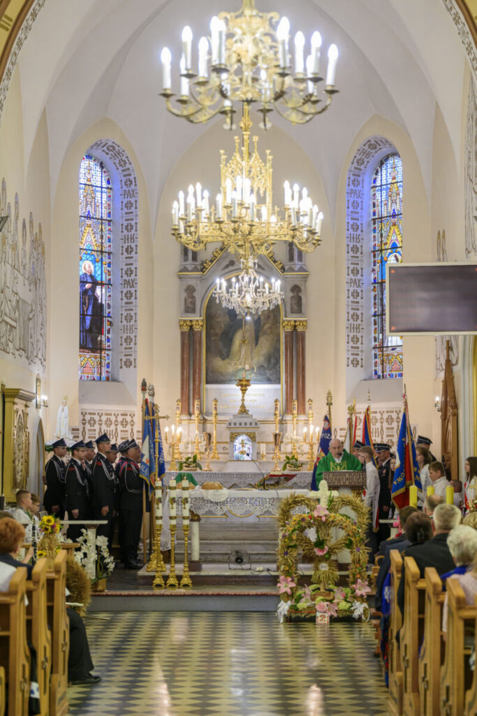 Wnętrze kościoła z ozdobnym ołtarzem i witrażami w tle. Jasne, złote żyrandole zwisają z sufitu. Ludzie w strojach galowych stoją przed ołtarzem.