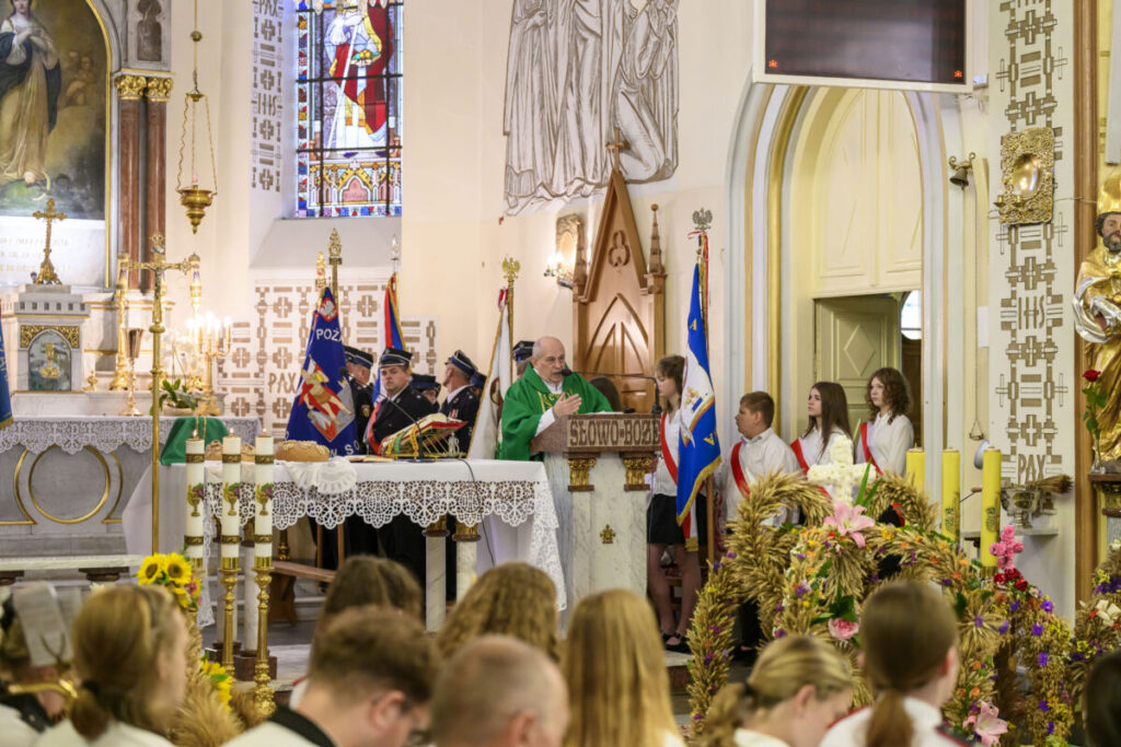 Kościół pełen wiernych, kapłan w zielonej szacie przy ambonie mówi kazanie. W tle kolorowe witraże i sztandary.