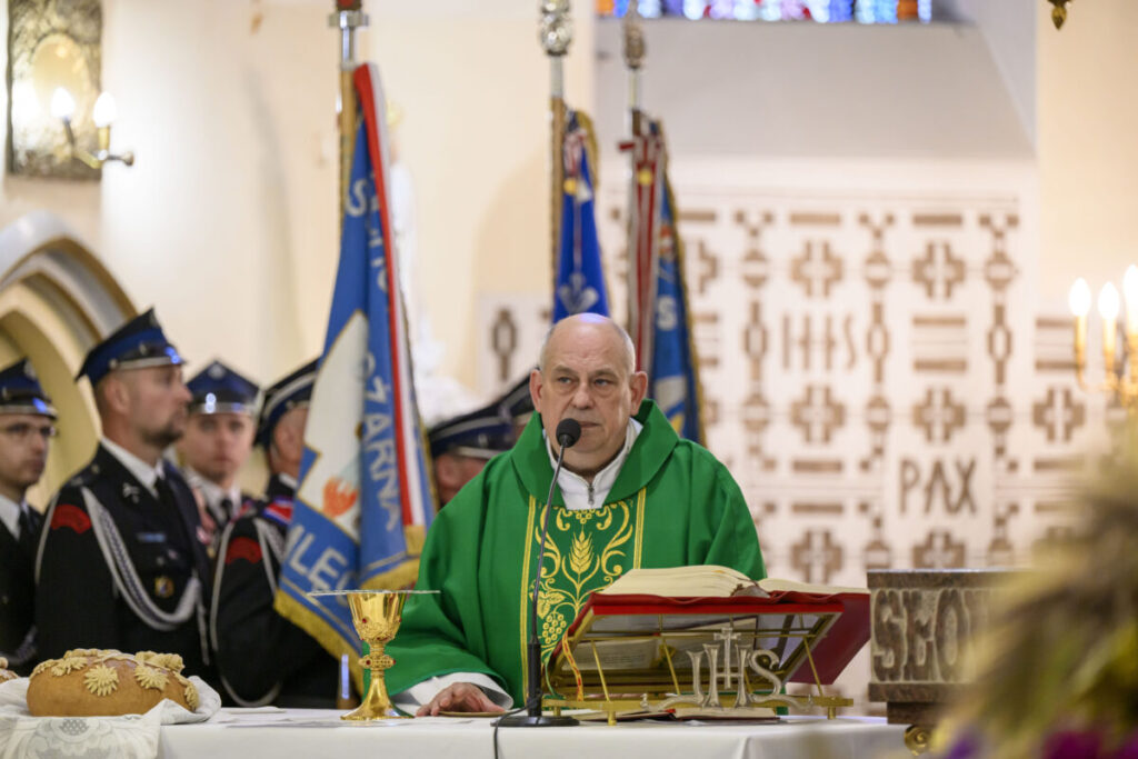 Kapłan w zielonym ornacie prowadzi mszę w kościele. Na pierwszym planie leży chleb i złoty kielich. W tle widoczni strażacy i sztandary.