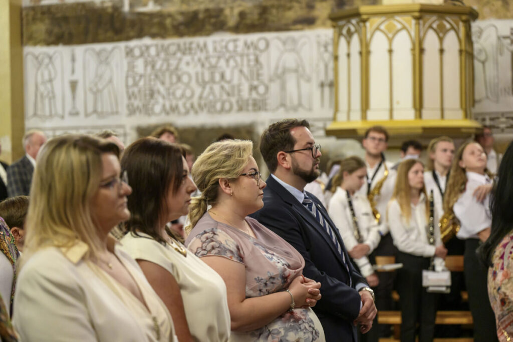 Grupa ludzi stoi w kościele, z przodu kilka osób w eleganckich strojach. W tle widoczna orkiestra młodzieżowa w białych koszulach. Ściany ozdobione freskami w jasnych tonacjach.