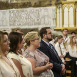 Grupa ludzi stoi w kościele, z przodu kilka osób w eleganckich strojach. W tle widoczna orkiestra młodzieżowa w białych koszulach. Ściany ozdobione freskami w jasnych tonacjach.