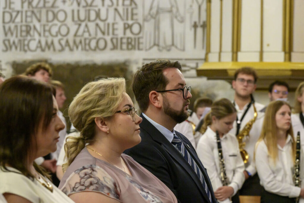 Grupa ludzi siedzi w kościele, w tle napis na ścianie. Dominują kolory białe i złote. Ludzie skupieni na wydarzeniu.