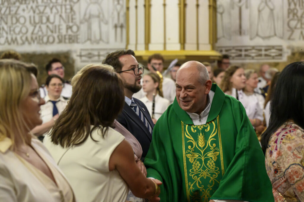 Na zdjęciu widać księdza w zielonej szacie liturgicznej, uśmiechającego się i witającego ludzi. Tło to wnętrze kościoła z ozdobionymi ścianami. Kolory dominujące to zieleń, biel i złoto.