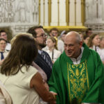 Na zdjęciu widać księdza w zielonej szacie liturgicznej, uśmiechającego się i witającego ludzi. Tło to wnętrze kościoła z ozdobionymi ścianami. Kolory dominujące to zieleń, biel i złoto.