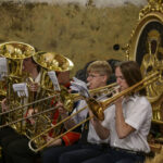 Grupa muzyków gra na puzonach w złotych odcieniach. W tle widoczny obraz z postaciami religijnymi. Dekoracje z kwiatami słonecznika.