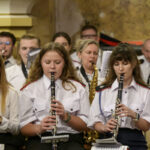 Na zdjęciu jest orkiestra dęta; trzy kobiety grają na klarnetach. Mają białe koszule, tło jest złotawo-brązowe. W tle widać innych muzyków i instrumenty.