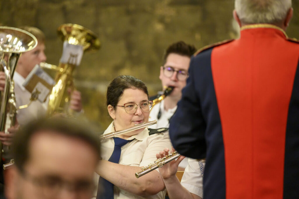 Osoby grające na instrumentach dętych w formalnych strojach. Dominują złote i czerwone kolory. Tło jest rozmyte i ciemne.