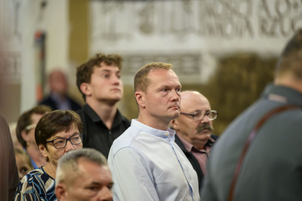 Na zdjęciu widać grupę osób stojących w pomieszczeniu. Dominują stonowane kolory ubrań i neutralne tło. Ludzie są skupieni, patrzą w jednym kierunku.