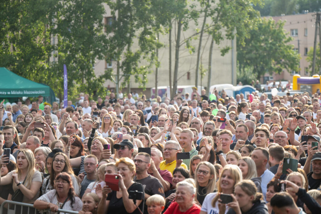 Zdjęcie przedstawia tłum ludzi na zewnątrz, w słoneczny dzień. W tle widoczne są drzewa i namiot zielonego koloru. Większość osób trzyma smartfony, wyglądają na rozradowane.