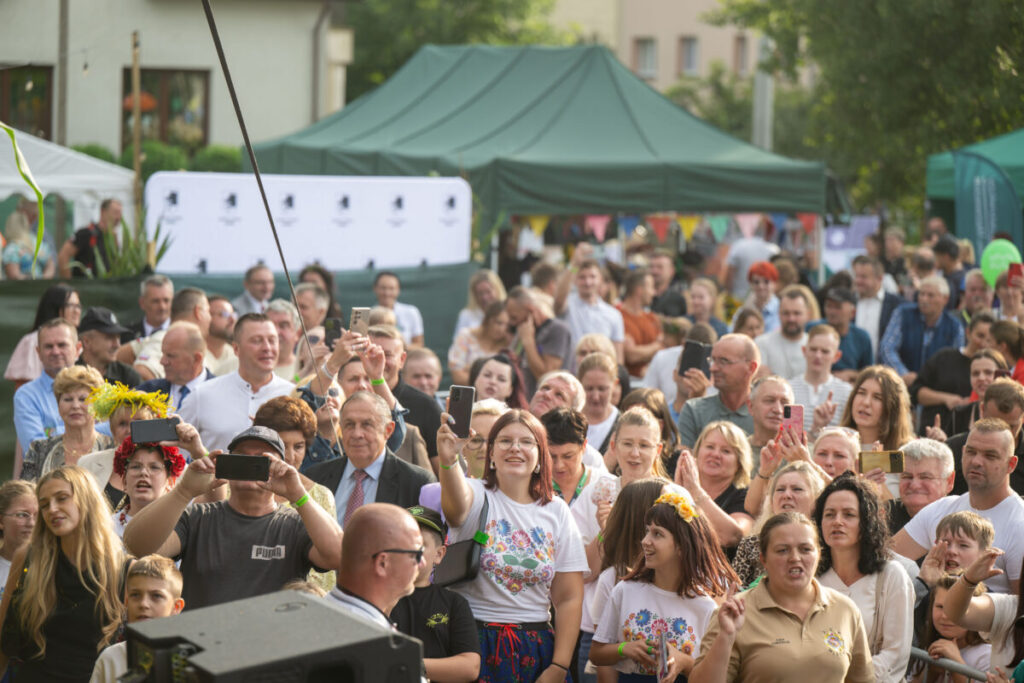 Grupa ludzi na plenerowym wydarzeniu, robiących zdjęcia telefonami. W tle zielone namioty i drzewa. Atmosfera radosna i słoneczna.